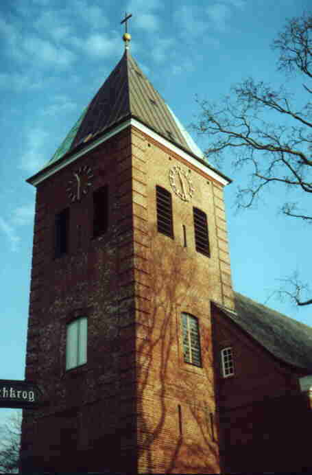 Ein Kirchturm aus braunen Steinen erbaut. Am unteren Bildrand sind Schießscharten zu erkennen. Oben die Schalllöcher der Glocken. Darüber das Ziffernblatt der Kirchturmuhr...