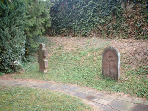 Foto: Zwei Gedenksteine an einem schmalen Weg: Ein Gedenkstein als russisch-orthodoxes Doppelkreuz und eine Gedenktafel mit einem Davidstern. Auf beiden Denkmälern liegen kleine Steine.