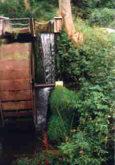 Links am Bildrand ein verrostetes Mühlrad, daneben fließt der Bach in einem kleinen Wasserfall herunter...